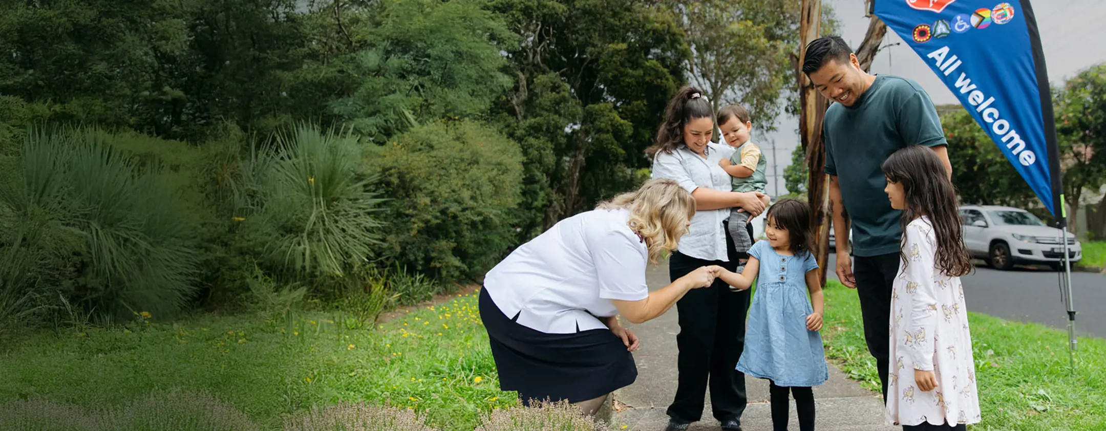 Female Salvos officer shaking hands with a girl and her family. The background is a path with surrounding grass and plants with a Salvos flag that says "All welcome".
