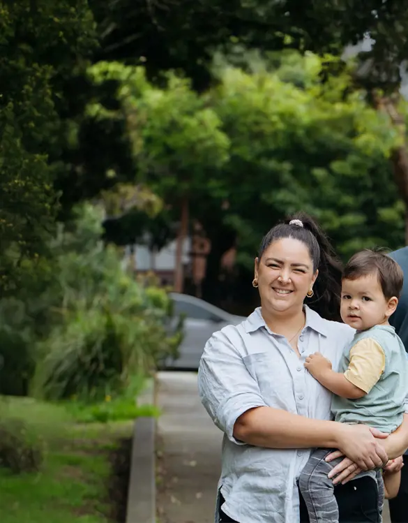Smiling young family of four enjoying a walk along a leafy suburban path — mum cradling the toddler while dad gently holds their daughter’s hand.