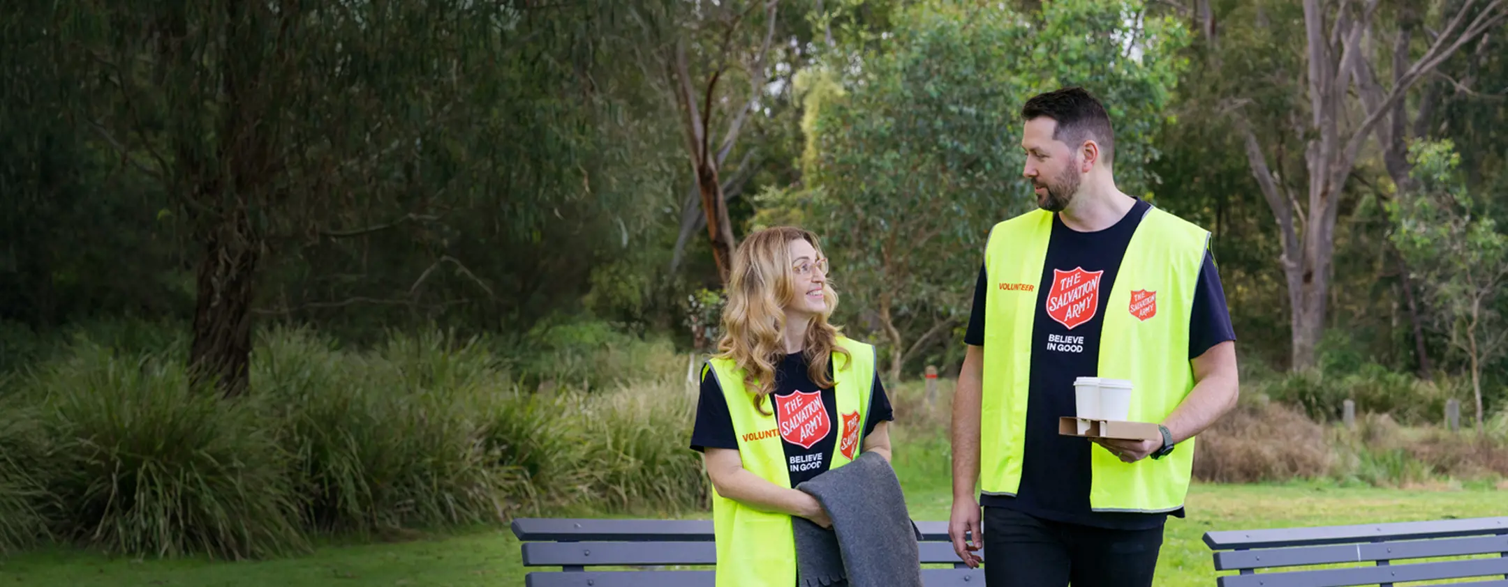 Two Salvation Army volunteers sharing a friendly conversation in a park, ready with warm coffee, blankets, and open hearts to support those doing it tough.