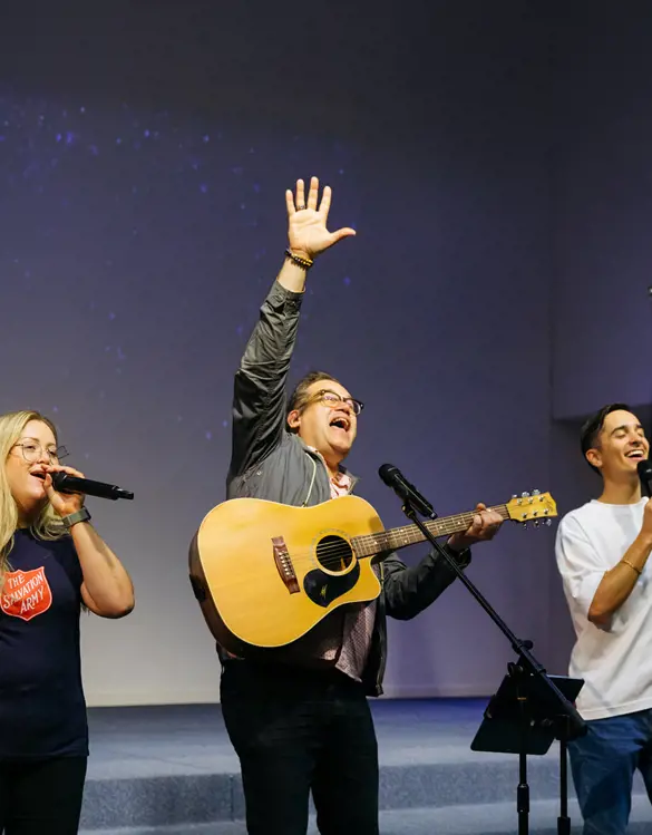 Three people leading worship on a church stage — one playing acoustic guitar, and the other 2 singing in a Salvation Army t-shirt, as heartfelt music fills the space.
