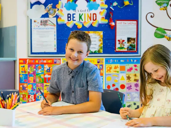 Two children sitting at a table smiling while drawing on paper.
