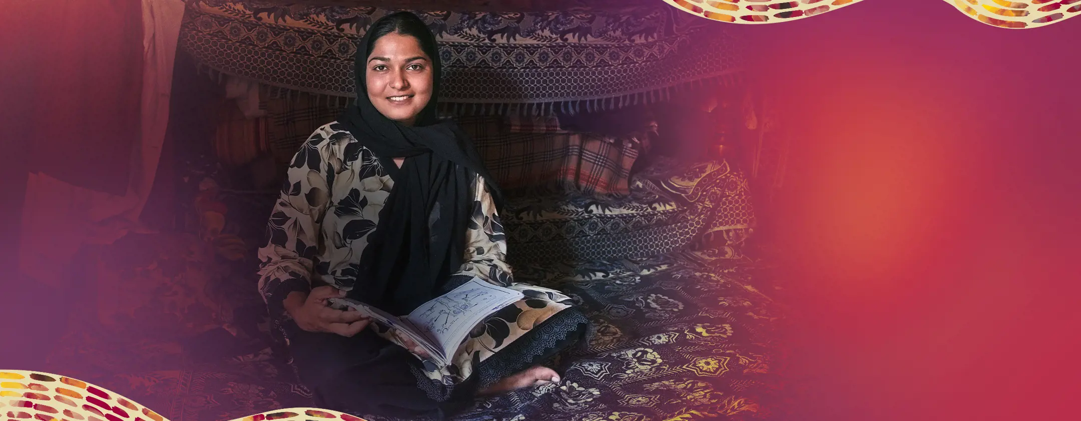 Woman smiling seated on patterned rugs inside a fabric-lined shelter, holding an open notebook and looking toward the camera.