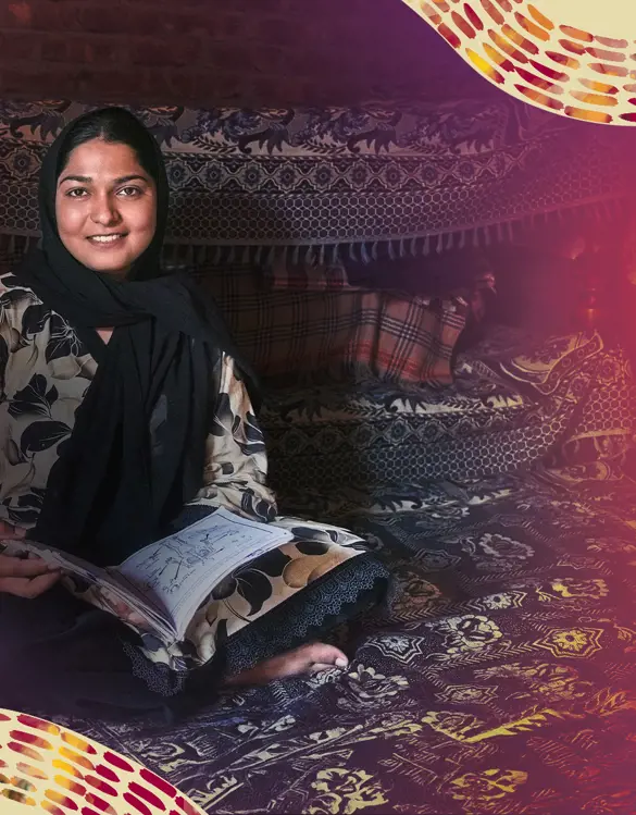 Woman smiling seated on patterned rugs inside a fabric-lined shelter, holding an open notebook and looking toward the camera.