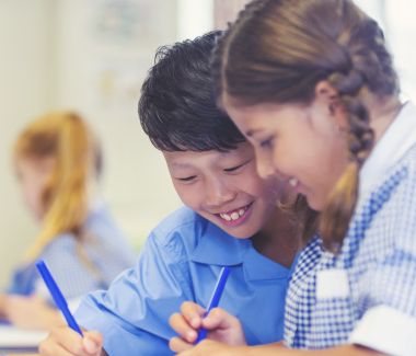 Two children are focused on writing in a classroom, surrounded by educational materials and a bright learning environment.