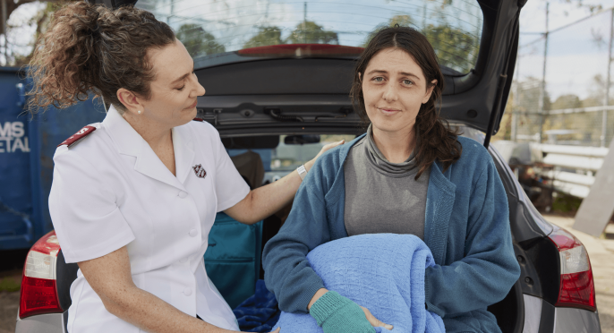 A Salvos Officer in a white uniform is holding a blanket next to a woman in blue, who is sitting in the trunk of a car.