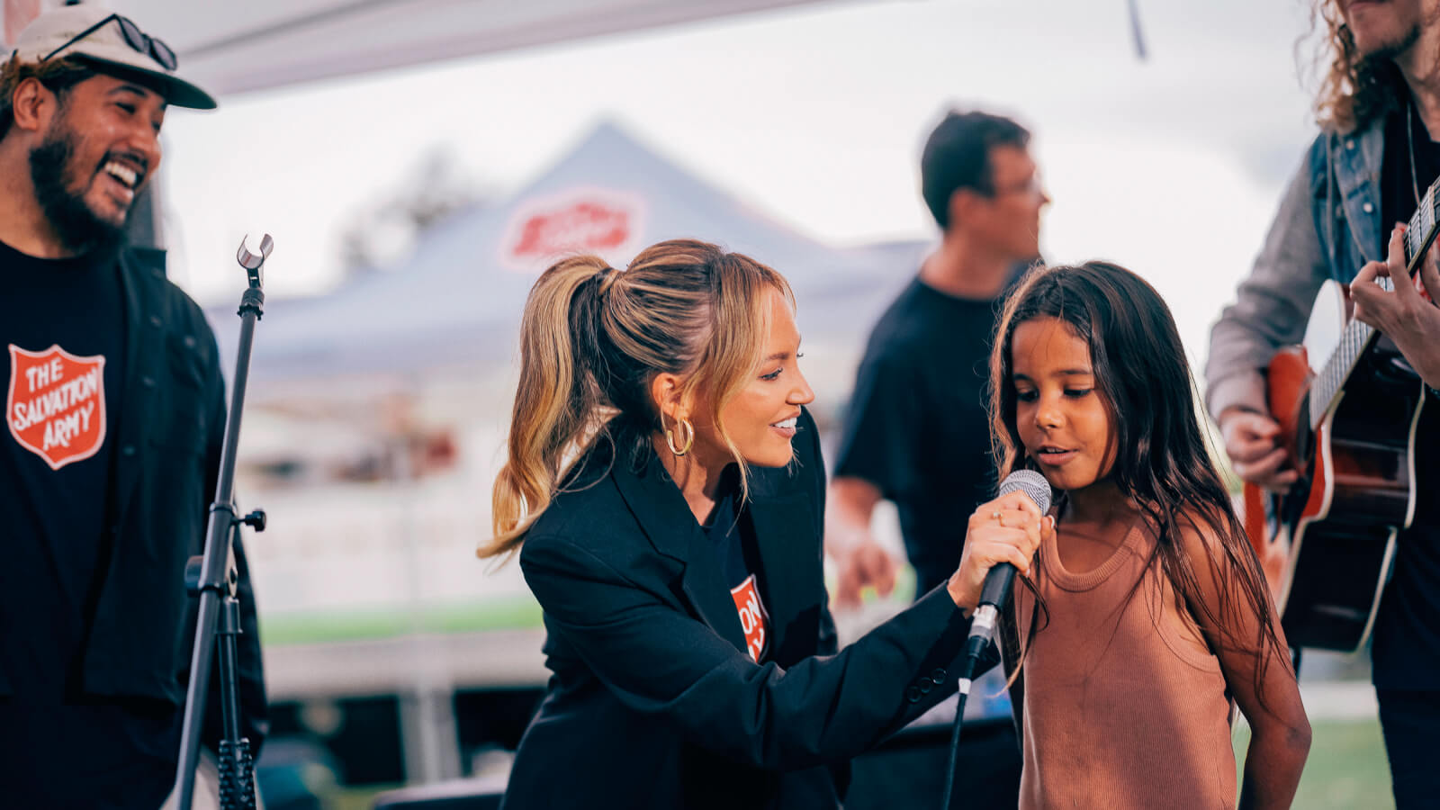 Samantha Jade, who is holding a microphone, is accompanied by a young girl, providing her with the opportunity to speak.