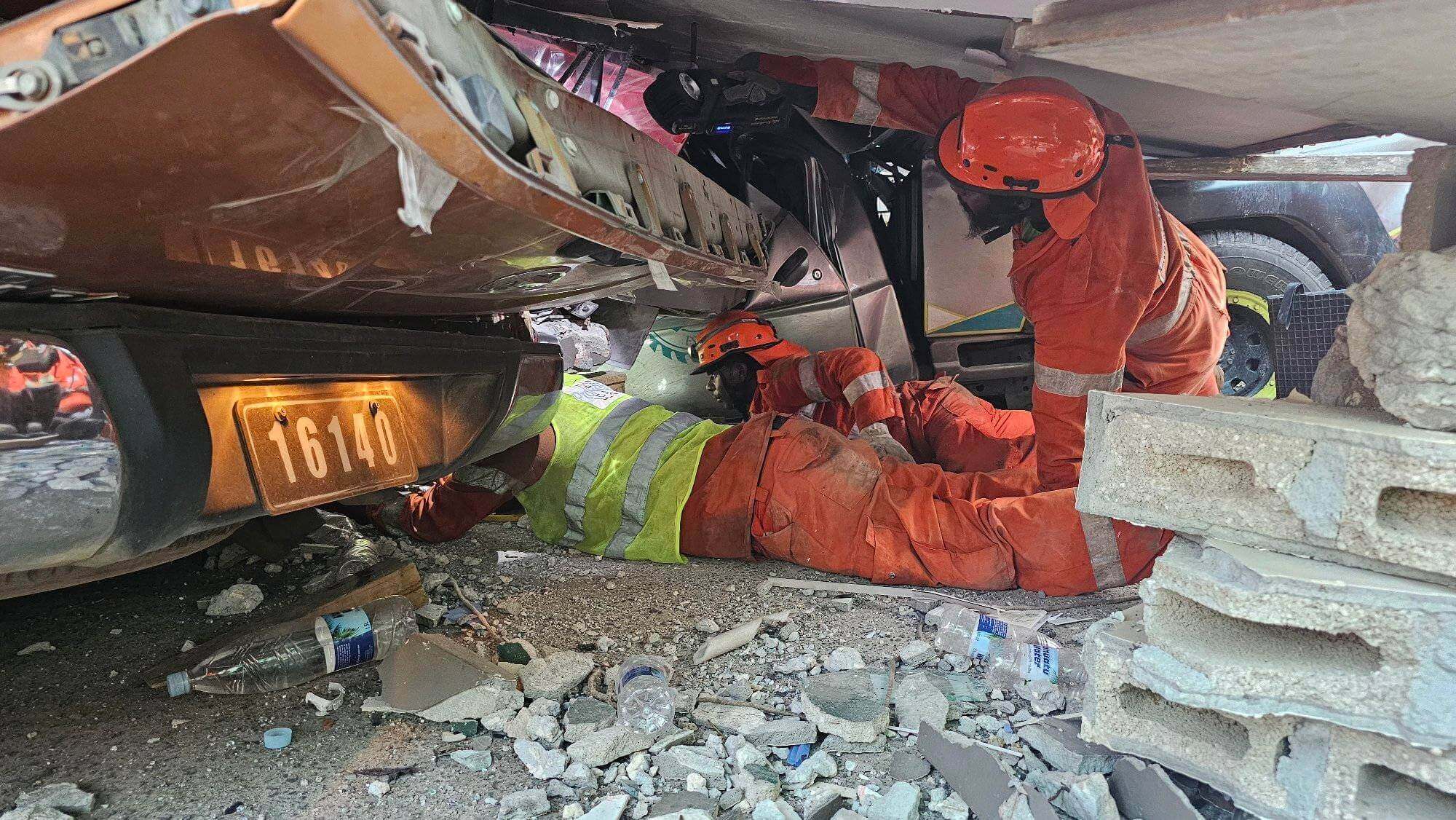 Rescue workers in orange suits and helmets crawl under debris and a vehicle. Rubble, bricks, and a plastic bottle are scattered around. 