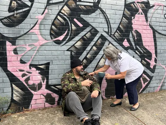 A Salvos officer offers a hot coffee to a man sitting against a graffiti-covered wall.