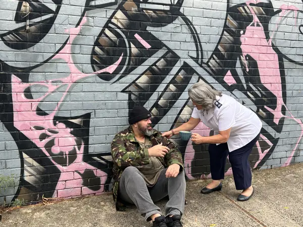 A Salvos officer offers a hot coffee to a man sitting against a graffiti-covered wall.